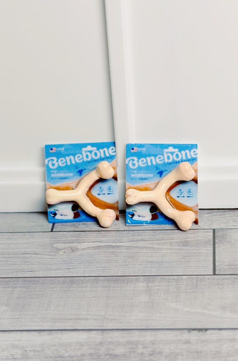 May include: Two Benebone wishbone dog chews, each in a blue package. The wishbones are tan and bone-shaped. The packaging has the brand name and a graphic. The items are displayed on a grey wood-effect surface.