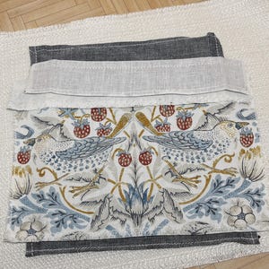 May include: Folded fabric with a detailed bird and strawberry design in blue, red, and gold on a white backdrop. It has a grey border and a light linen-like trim. The fabric rests on a textured white surface.