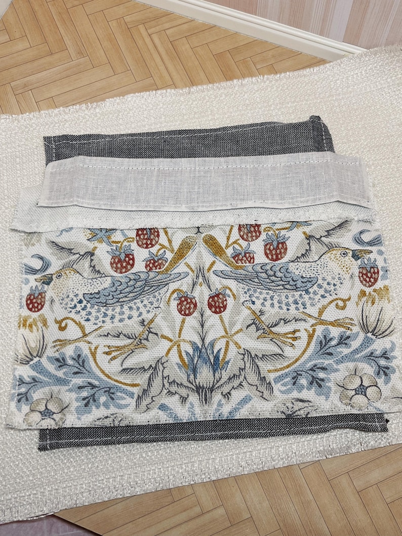 May include: Folded fabric with a detailed bird and strawberry design in blue, red, and gold on a white backdrop. It has a grey border and a light linen-like trim. The fabric rests on a textured white surface.