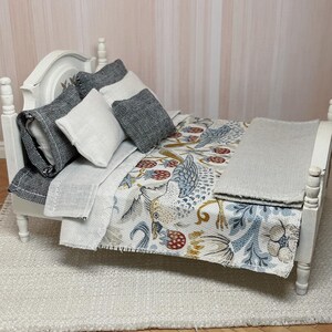 May include: A miniature white bed with a headboard, featuring pillows and a patterned quilt. The quilt displays birds, flowers, and berries in blue, yellow, and red. A grey blanket is neatly placed at the foot of the bed.