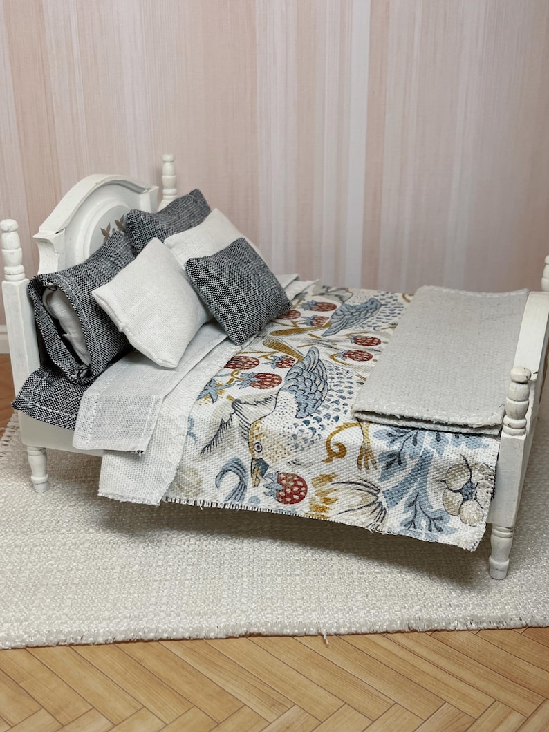 May include: A miniature white bed with a headboard, featuring pillows and a patterned quilt. The quilt displays birds, flowers, and berries in blue, yellow, and red. A grey blanket is neatly placed at the foot of the bed.