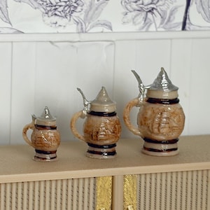 May include: Three miniature ceramic beer steins with silver-colored lids and handles. The steins are brown with a textured design and dark brown bands. They are arranged on a light-colored surface.