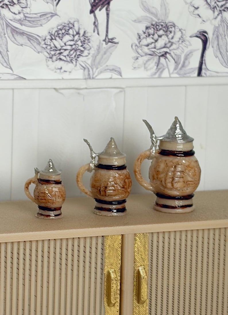May include: Three miniature ceramic beer steins with silver-colored lids and handles. The steins are brown with a textured design and dark brown bands. They are arranged on a light-colored surface.