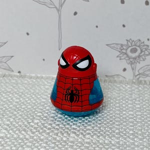 May include: A red, blue, and black Spider-Man figurine. The figure has a red head with white eyes and a black spider emblem on the chest. The body is blue with a red web design.
