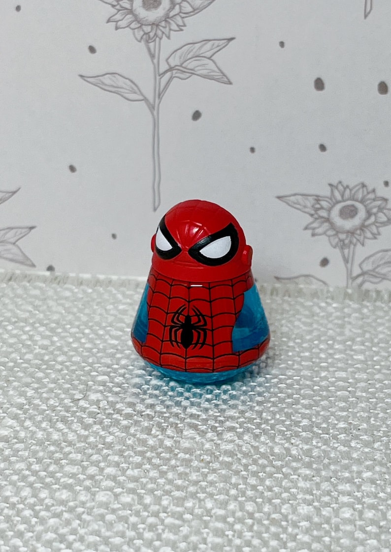 May include: A red, blue, and black Spider-Man figurine. The figure has a red head with white eyes and a black spider emblem on the chest. The body is blue with a red web design.