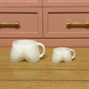 May include: Two white ceramic mugs shaped like a pair of shorts. One mug is larger than the other. The mugs have a handle and are sitting on a wooden surface. The background features a pink cabinet.