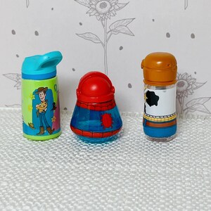 May include: Three colorful, miniature character-themed bottles. The first is a blue and green water bottle with a Woody cartoon design. The second is a red and blue container. The third is a bottle with a brown top and a black and white cow print.