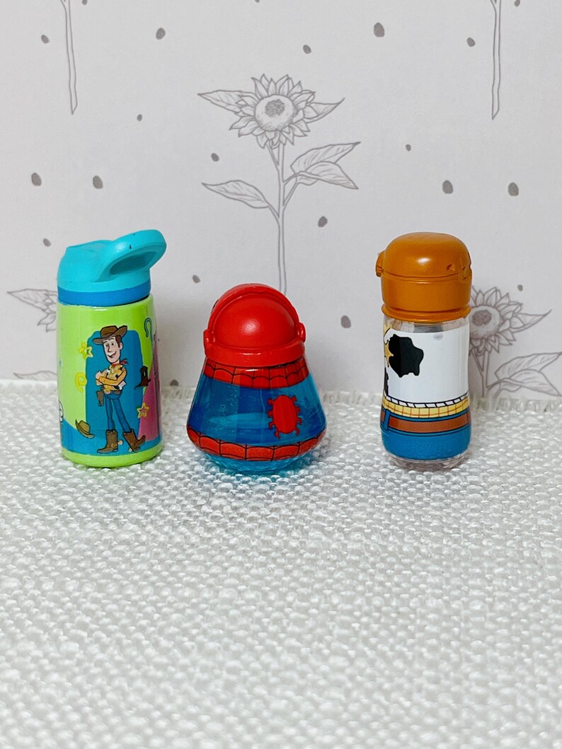 May include: Three colorful, miniature character-themed bottles. The first is a blue and green water bottle with a Woody cartoon design. The second is a red and blue container. The third is a bottle with a brown top and a black and white cow print.
