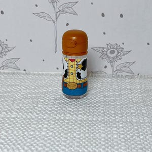 May include: A small, clear plastic container with a brown cap. The container is decorated with a cartoon image of a character wearing a yellow shirt, a brown vest, and a blue bottom. The container is standing on a white surface.