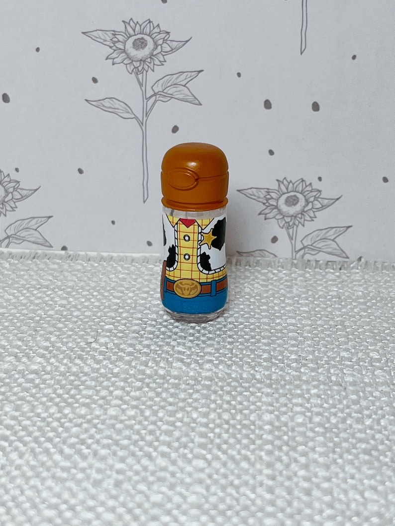 May include: A small, clear plastic container with a brown cap. The container is decorated with a cartoon image of a character wearing a yellow shirt, a brown vest, and a blue bottom. The container is standing on a white surface.