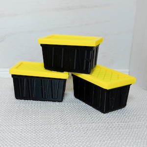 May include: Three small, black storage bins with yellow lids are stacked on a white surface. The bins have a rectangular shape with vertical grooves on the sides. The lids are a bright yellow color.