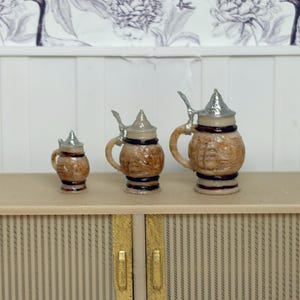 May include: Three miniature beer steins with silver lids and handles. The steins are brown with decorative relief patterns and sit on a light-colored cabinet. The background features a floral wallpaper.