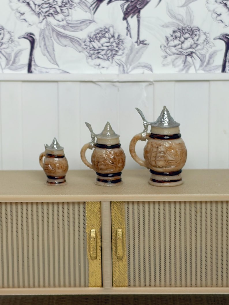 May include: Three miniature beer steins with silver lids and handles. The steins are brown with decorative relief patterns and sit on a light-colored cabinet. The background features a floral wallpaper.