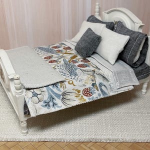 May include: A miniature white bed with a headboard and footboard. The bed is made up with a patterned quilt featuring birds and berries in blue, red, and yellow. Several pillows in shades of grey and white are arranged on the bed. A grey blanket is folded at the foot of the bed.