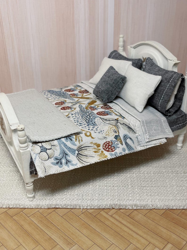 May include: A miniature white bed with a headboard and footboard. The bed is made up with a patterned quilt featuring birds and berries in blue, red, and yellow. Several pillows in shades of grey and white are arranged on the bed. A grey blanket is folded at the foot of the bed.