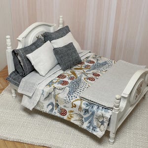 May include: A miniature white bed with a patterned comforter featuring birds and berries, and grey and white pillows. The bed is on a white rug, creating a cosy scene. The bed frame has decorative carvings.