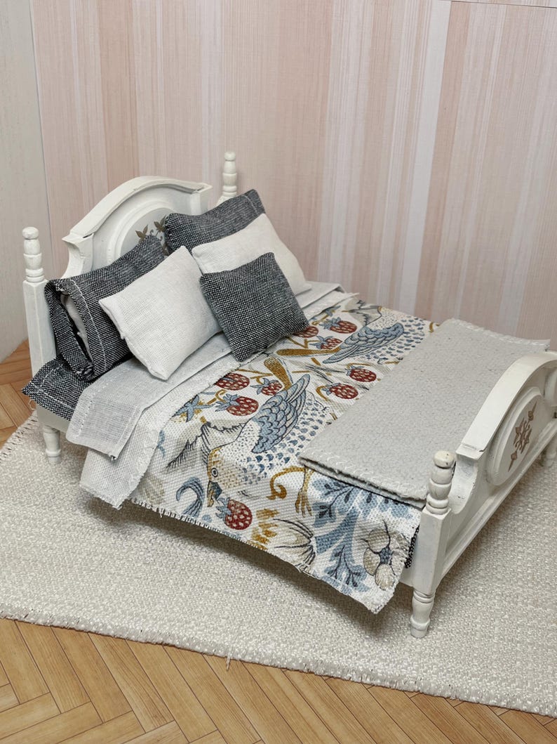 May include: A miniature white bed with a patterned comforter featuring birds and berries, and grey and white pillows. The bed is on a white rug, creating a cosy scene. The bed frame has decorative carvings.