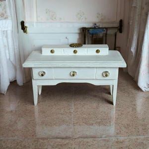 May include: A miniature white wooden table with three drawers and crystal knobs. The table has a matching rectangular top piece with three gold knobs. A small gold-colored decorative item sits on top. The table is in a dollhouse setting.