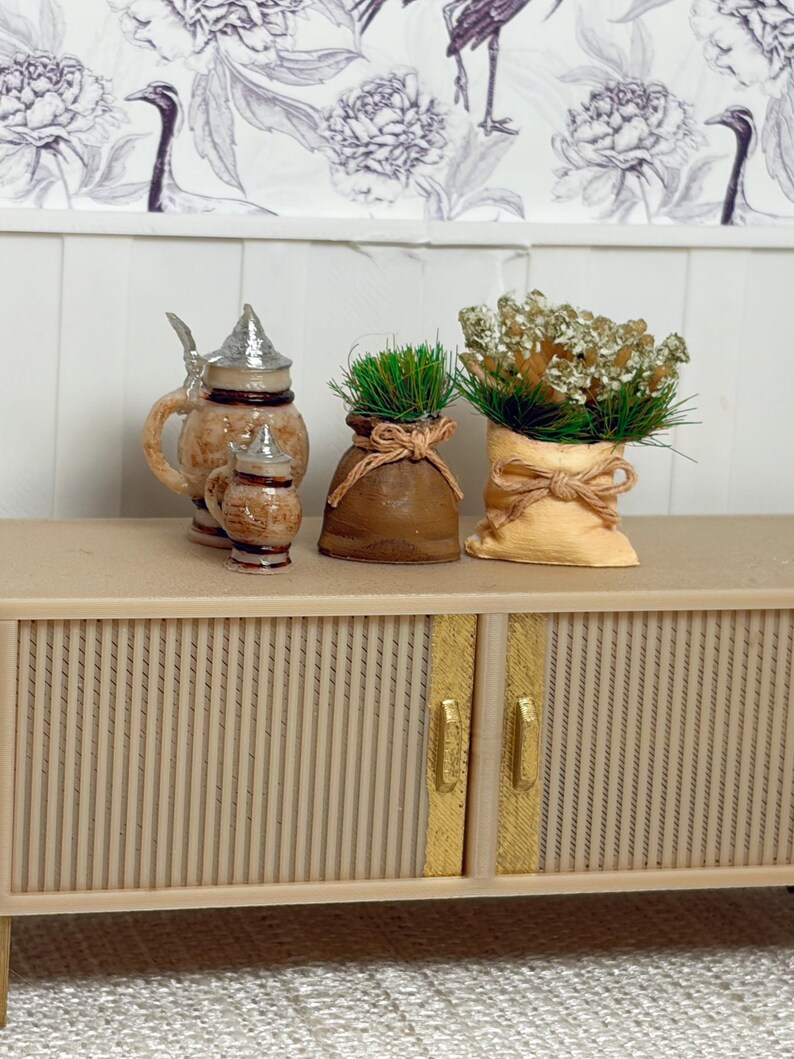 May include: Miniature decorative items on a beige cabinet. Includes a small beer stein set with silver lids, two small burlap sacks tied with twine, one with green grass and the other with dried flowers. Floral wallpaper in the background.