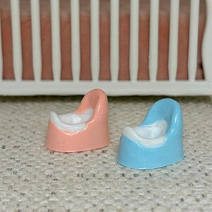May include: Miniature pink and blue potty chairs with white interiors. The small chairs are in front of a white crib with a pink mattress. The potty chairs are likely for a dollhouse or other miniature setting.