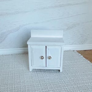 May include: A miniature white cabinet with two doors and crystal-like knobs. The small cabinet has a rectangular shape and a small backsplash. The cabinet is placed on a light gray woven rug.