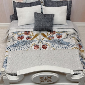May include: A miniature white bed with a patterned quilt featuring birds and strawberries. The bed is dressed with white and grey pillows, creating a cosy scene. The bed frame has a decorative floral design. The quilt and pillows are in a miniature scale.