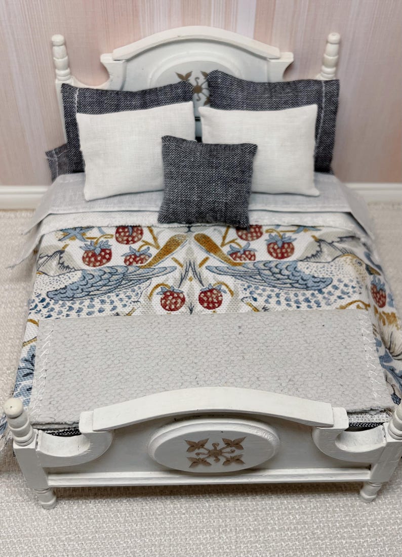 May include: A miniature white bed with a patterned quilt featuring birds and strawberries. The bed is dressed with white and grey pillows, creating a cosy scene. The bed frame has a decorative floral design. The quilt and pillows are in a miniature scale.