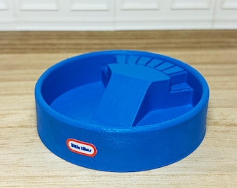 May include: A bright blue Little Tikes toy pool with a built-in slide and steps. The pool is round with a raised edge and a small Little Tikes logo on the side. The toy is made of plastic and sits on a wooden surface.