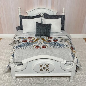 May include: A miniature white bed with a decorative headboard and footboard. The bed is made up with a patterned comforter featuring birds and berries, white and grey pillows. The bed is set on a grey rug.