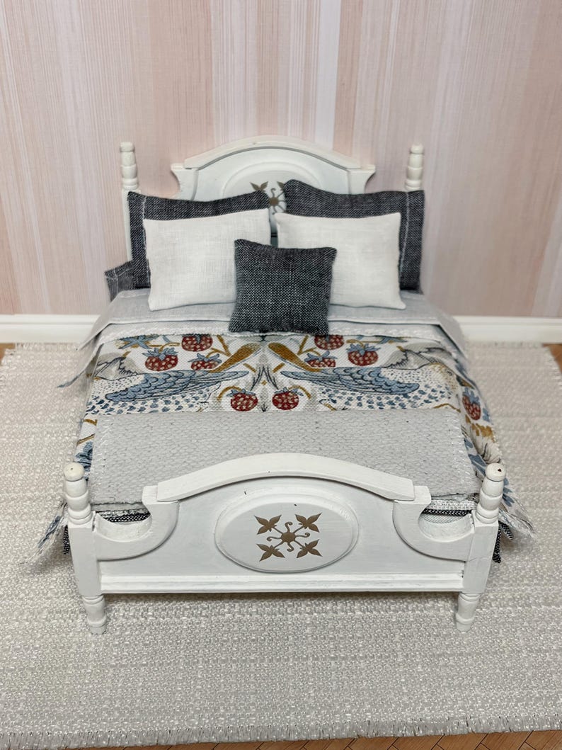 May include: A miniature white bed with a decorative headboard and footboard. The bed is made up with a patterned comforter featuring birds and berries, white and grey pillows. The bed is set on a grey rug.