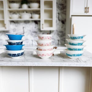 May include: A collection of vintage-style ceramic bowls in various colors and patterns. The bowls are stacked on a white countertop. The colors include blue, pink, and turquoise, with floral and geometric designs. The kitchen setting includes white cabinets and a patterned wallpaper.