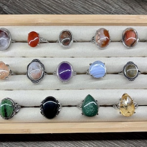 May include: A wooden display case with 16 rings featuring various oval and teardrop shaped gemstones. The rings are made of silver and feature a variety of colors, including green, blue, purple, orange, and brown.
