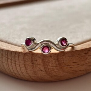 May include: A silver ring with a wavy design, featuring three round, vibrant pink gemstones. The ring is set against a light-colored background, showcasing its elegant and minimalist style. The gemstones are approximately 0.12 inches in diameter.