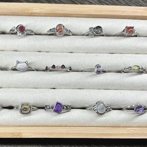 May include: A wooden box with a white velvet interior displays a collection of 18 silver rings with various gemstones. The rings are arranged in rows, showcasing a variety of colours and styles.