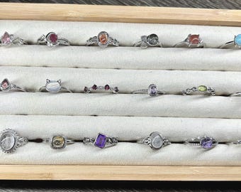May include: A wooden box with a white velvet interior displays a collection of 18 silver rings with various gemstones. The rings are arranged in rows, showcasing a variety of colors and styles.
