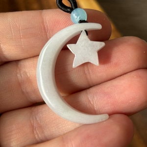 Puede incluir: Pendiente de jade blanco en forma de media luna con un dije de estrella de jade blanco. El pendiente está encordado en un cordón elástico negro con una cuenta de jade azul claro.