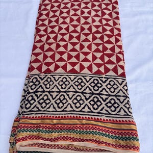 May include: Folded fabric with a geometric pattern in red and beige. The fabric has a border with black, gold, green, and red designs. The fabric is likely cotton or a similar material.