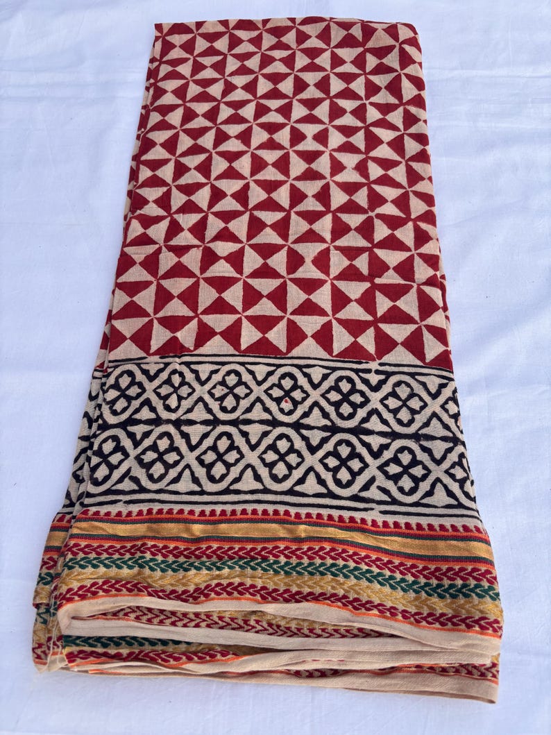 May include: Folded fabric with a geometric pattern in red and beige. The fabric has a border with black, gold, green, and red designs. The fabric is likely cotton or a similar material.