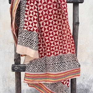May include: A draped textile with a geometric pattern of red triangles on a cream background. The border features black and white geometric shapes with red, yellow, and green stripes. The fabric is displayed on a wooden ladder.