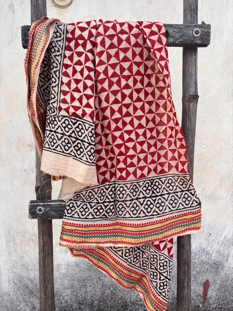 May include: A draped textile with a geometric pattern of red triangles on a cream background. The border features black and white geometric shapes with red, yellow, and green stripes. The fabric is displayed on a wooden ladder.