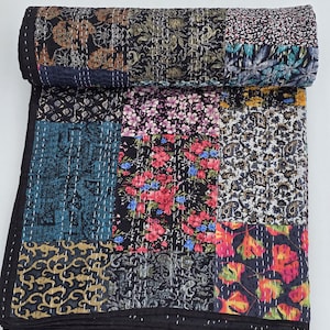 Könnte beinhalten: Ein Patchwork-Quilt mit schwarzem Rand, der verschiedene quadratische Stoffe mit Blumenmustern in Blau-, Rot- und Goldtönen aufweist. Der Quilt ist mit weißem Faden handgenäht und oben aufgerollt.