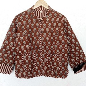 Pode incluir: Casaco acolchoado castanho com padrão floral. O casaco tem gola mandarim, mangas compridas e dois bolsos frontais. Os punhos e a gola interior apresentam um design listrado. O casaco está pendurado num cabide de madeira.