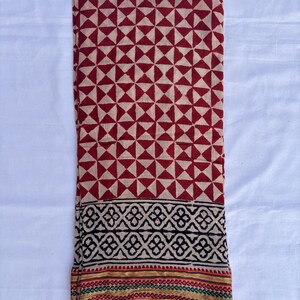 May include: A folded textile featuring a geometric pattern of red triangles on a beige background. The bottom border has a black and beige design with a thin stripe of red, green, and gold. The fabric is likely cotton or a similar material.
