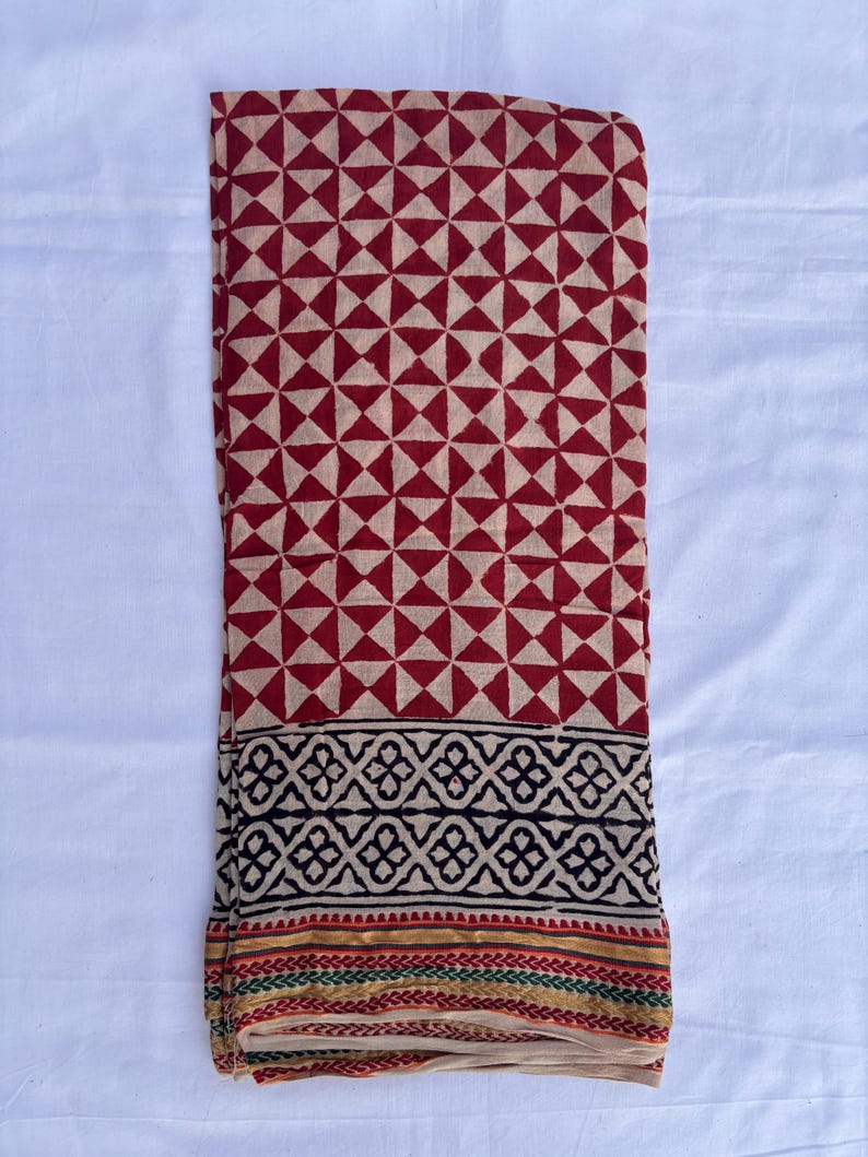 May include: A folded textile featuring a geometric pattern of red triangles on a beige background. The bottom border has a black and beige design with a thin stripe of red, green, and gold. The fabric is likely cotton or a similar material.