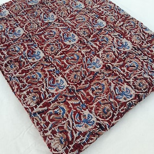 May include: A burgundy quilt with a floral pattern in blue, tan, and white. The quilt is made of a soft fabric and has a stitched design. The quilt is folded and placed on a white surface.