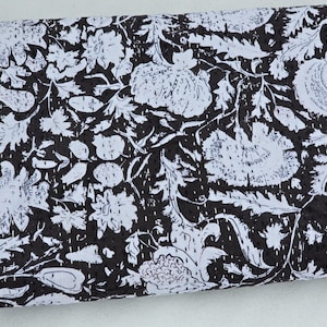 May include: A folded textile with a black and white floral pattern. The design features large, stylized flowers and leaves against a dark background. The fabric appears to be quilted, with visible stitching throughout the design. The overall aesthetic is a blend of classic and contemporary styles.