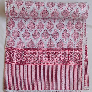 May include: A rolled-up white and red quilt with a repeating pattern of stylized teardrop shapes. The quilt features a border with geometric designs and vertical stripes. The fabric is soft and appears to be cotton.
