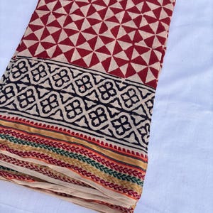 May include: Folded fabric with a geometric pattern. The top section features a repeating pattern of red triangles on a beige background. A black and beige border with a floral design is at the bottom, with colorful stripes.