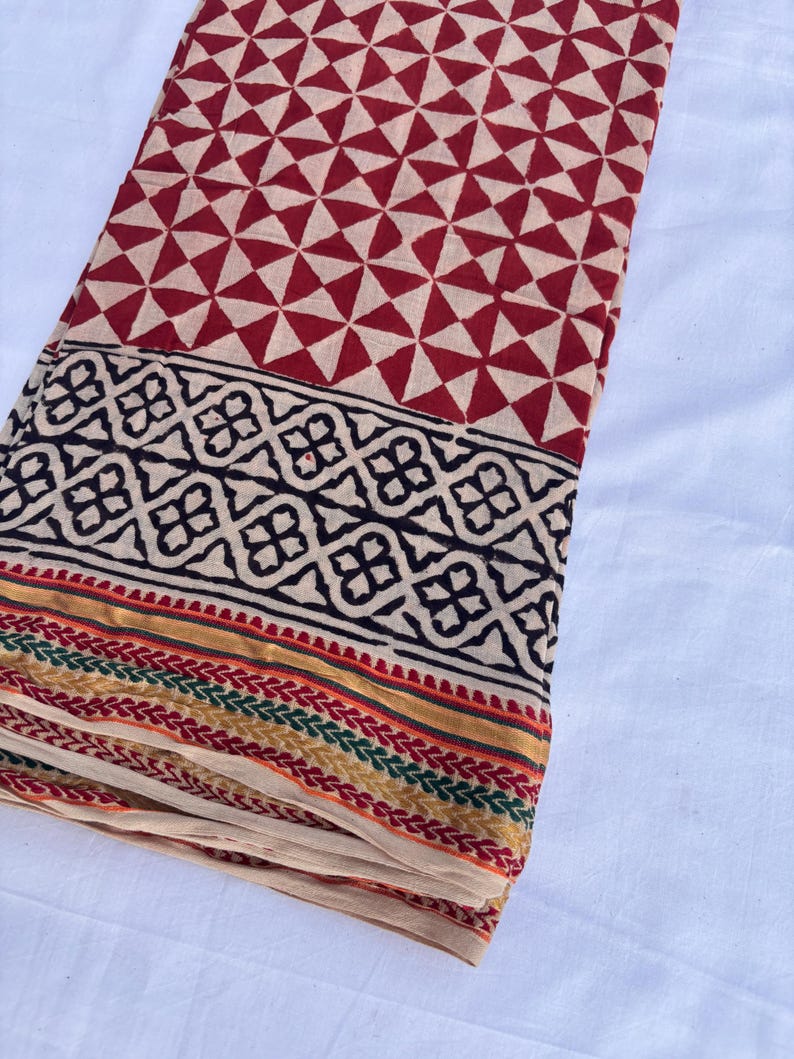 May include: Folded fabric with a geometric pattern. The top section features a repeating pattern of red triangles on a beige background. A black and beige border with a floral design is at the bottom, with colorful stripes.