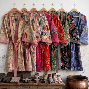 May include: A row of seven cotton robes hanging on wooden hangers, each featuring distinct floral block-print patterns in various colors including yellow, pink, red, green, black, and blue, with striped trim on the collars and matching waist ties.